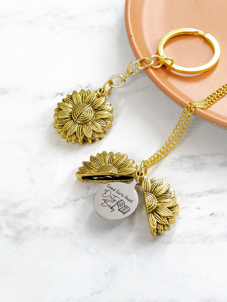 May include: A gold-colored sunflower pendant necklace and keychain. The sunflower pendant opens to reveal a silver charm with the text "Good Girls Read Dirty Books" and a book graphic. The keychain has a gold ring.