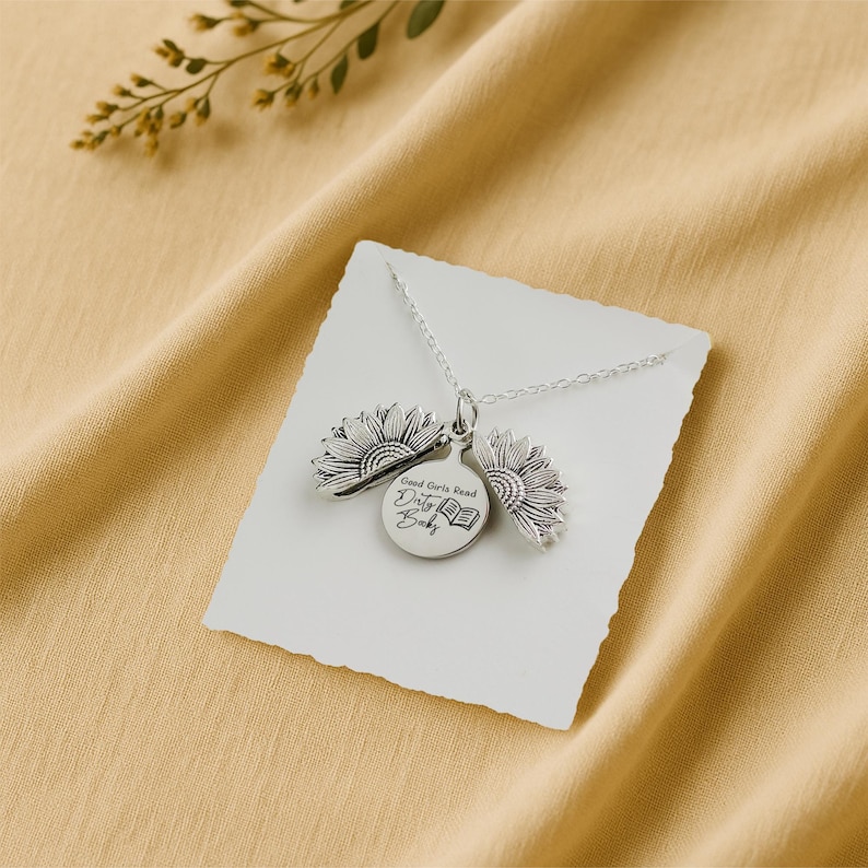May include: A silver necklace with a sunflower pendant. The pendant has a round charm with the text "Good Girls Read Dirty Books" and an image of a book. The necklace is displayed on a white card against a yellow background.