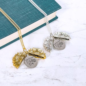 May include: Two sunflower pendant necklaces, one gold-toned and one silver-toned, with a chain. Each pendant opens to reveal a disc engraved with "Good Girls Read Dirty Books" and a book graphic. The necklaces rest on a marble surface.