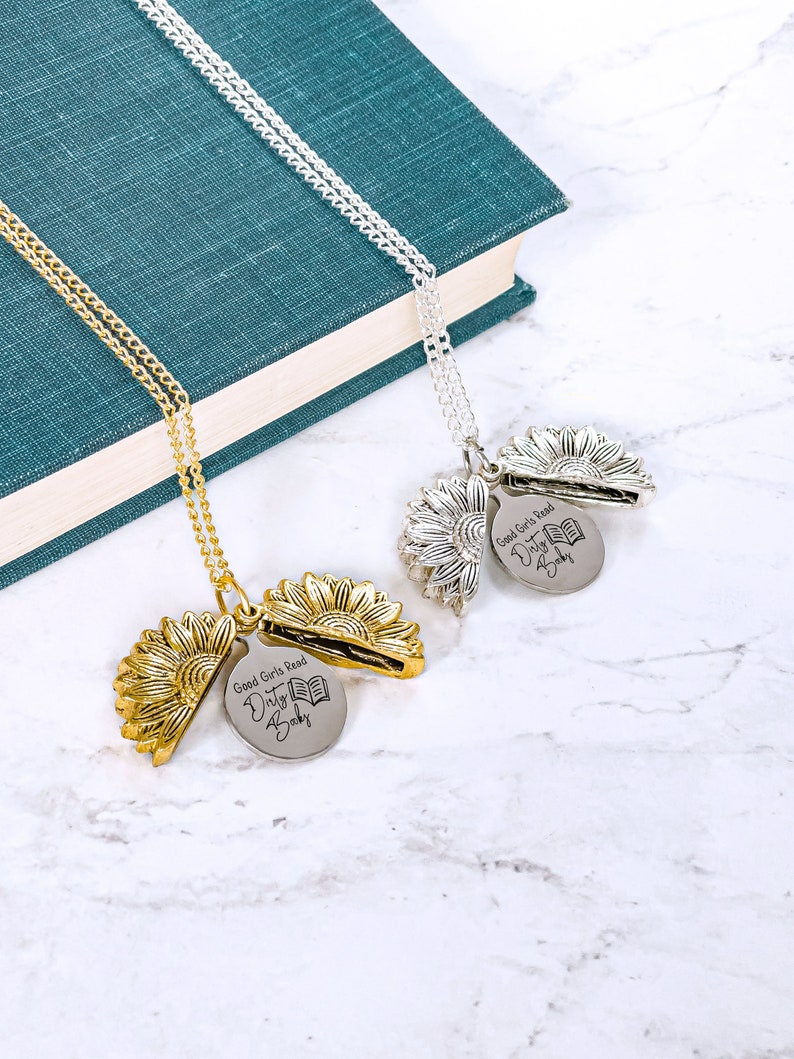May include: Two sunflower pendant necklaces, one gold-toned and one silver-toned, with a chain. Each pendant opens to reveal a disc engraved with "Good Girls Read Dirty Books" and a book graphic. The necklaces rest on a marble surface.