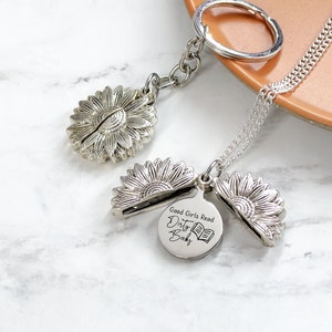May include: A silver-colored sunflower keychain and necklace set. The keychain features a sunflower charm, and the necklace has a sunflower pendant with a round charm that reads "Good Girls Read Dirty Books" and a book graphic.