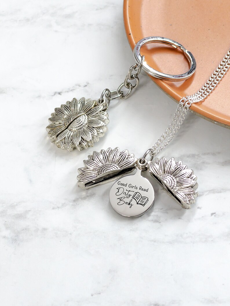 May include: A silver-colored sunflower keychain and necklace set. The keychain features a sunflower charm, and the necklace has a sunflower pendant with a round charm that reads "Good Girls Read Dirty Books" and a book graphic.
