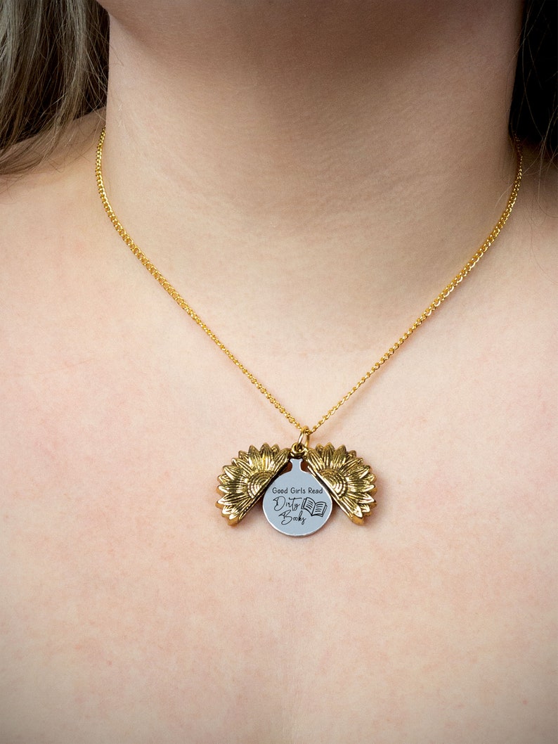 May include: A gold-colored necklace with a sunflower pendant. The pendant opens to reveal a circular charm with the text "Good Girls Read Dirty Books" and a book graphic. The necklace is worn around the neck.