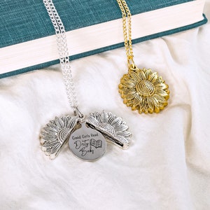 May include: Two sunflower pendant necklaces, one silver and one gold, rest on a white surface. The silver necklace has a pendant that opens to reveal a disc with the text "Good Girls Read Dirty Books" and a book graphic.
