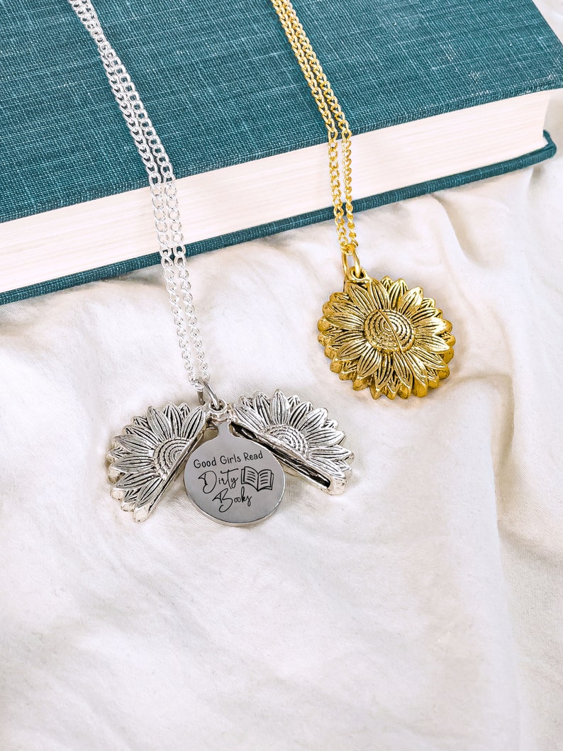 May include: Two sunflower pendant necklaces, one silver and one gold, rest on a white surface. The silver necklace has a pendant that opens to reveal a disc with the text "Good Girls Read Dirty Books" and a book graphic.