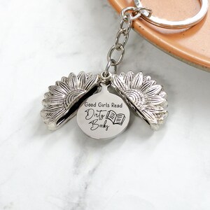 May include: Silver-colored keychain featuring a sunflower design that opens to reveal a circular charm. The charm has the text "Good Girls Read Dirty Books" and an illustration of an open book. The keychain is attached to a silver keyring.