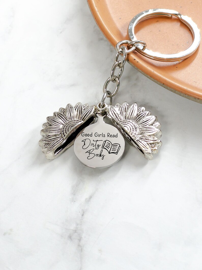 May include: Silver-colored keychain featuring a sunflower design that opens to reveal a circular charm. The charm has the text "Good Girls Read Dirty Books" and an illustration of an open book. The keychain is attached to a silver keyring.