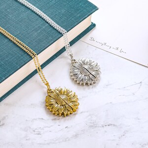 May include: Two sunflower pendant necklaces, one gold-toned and one silver-toned, rest on a marble surface. The gold necklace is on the left, and the silver necklace is on the right. The necklaces are on a teal book and a white card with the text "Do everything with Love."