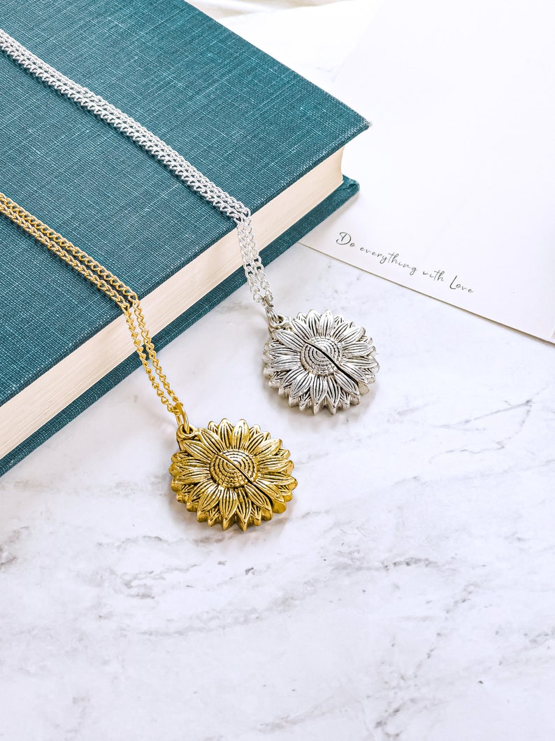 May include: Two sunflower pendant necklaces, one gold-toned and one silver-toned, rest on a marble surface. The gold necklace is on the left, and the silver necklace is on the right. The necklaces are on a teal book and a white card with the text "Do everything with Love."