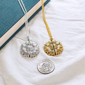 May include: Two sunflower pendant necklaces, one silver-toned and one gold-toned, are displayed. The pendants are open, revealing the inner details. A U.S. quarter coin is placed nearby for size comparison. The necklaces rest on a white fabric.