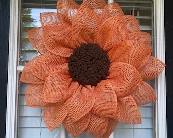 May include: A large, orange burlap sunflower wreath with a dark brown center, hanging on a white-framed window. The wreath's petals are layered, creating a dimensional effect. The window has white blinds.