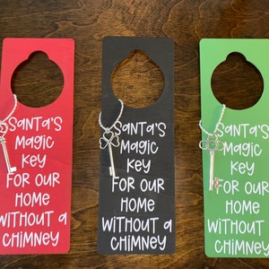 May include: Three wooden door hangers with a keyhole and a silver key charm. Each hanger has a different color: red, black, and green. The text on each hanger reads: "Santa's Magic Key For Our Home Without a Chimney".