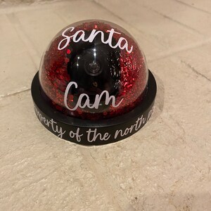 May include: A black and red Santa Cam decoration. The clear dome is filled with red glitter and features the words "Santa Cam" in white script. The base is black with the words "property of the north pole" in white.