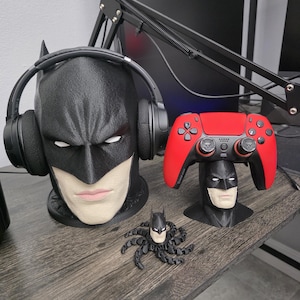 May include: A black Batman-themed headphone stand with headphones, a red and black game controller stand, and a small Batman-shaped octopus figurine on a wooden surface. The headphone stand has a detailed face.