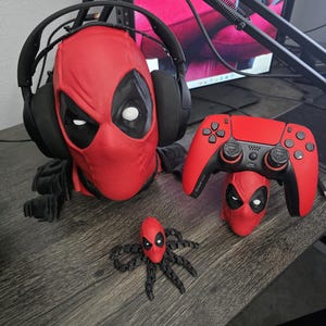 May include: A red Deadpool mask with black eye cutouts holds a black headset. A red PlayStation controller and a Deadpool-themed spider-like figure are also on the desk. The items are on a dark wood-grain surface.