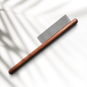 May include: A wooden comb with fine metal teeth, designed for grooming. The comb has a dark brown wooden handle and a rectangular head with closely spaced, silver-coloured teeth. The comb is set against a white background with shadows.