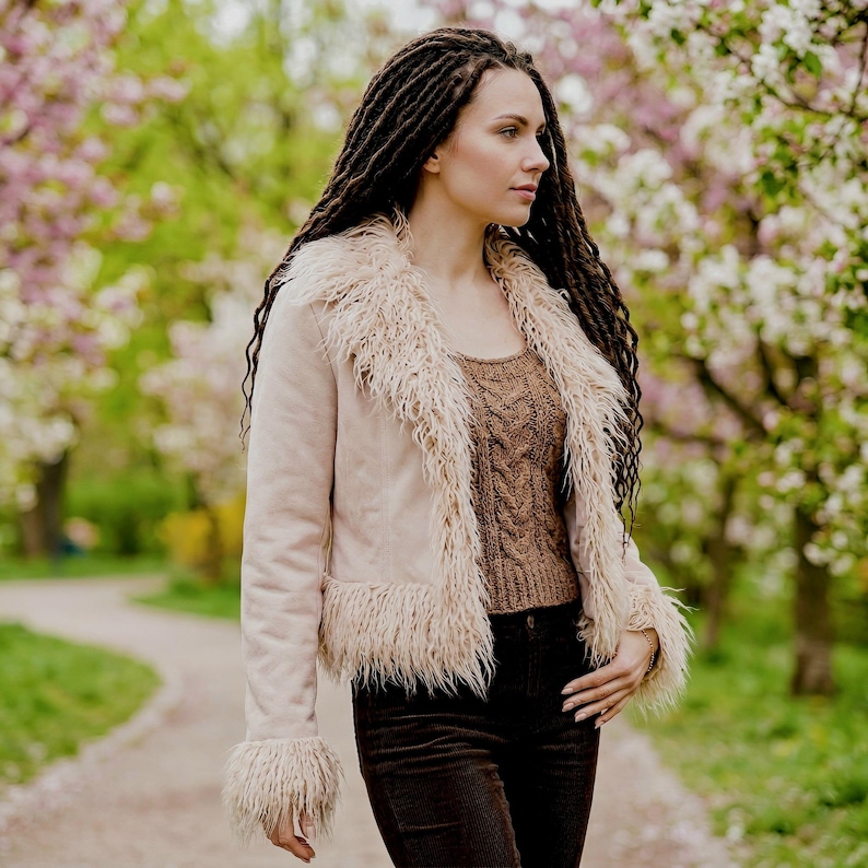 May include: A light pink jacket with faux fur trim around the collar, cuffs, and hem. The jacket is worn over a brown knit sweater and paired with dark brown corduroy pants. The background features a blurred path and flowering trees.