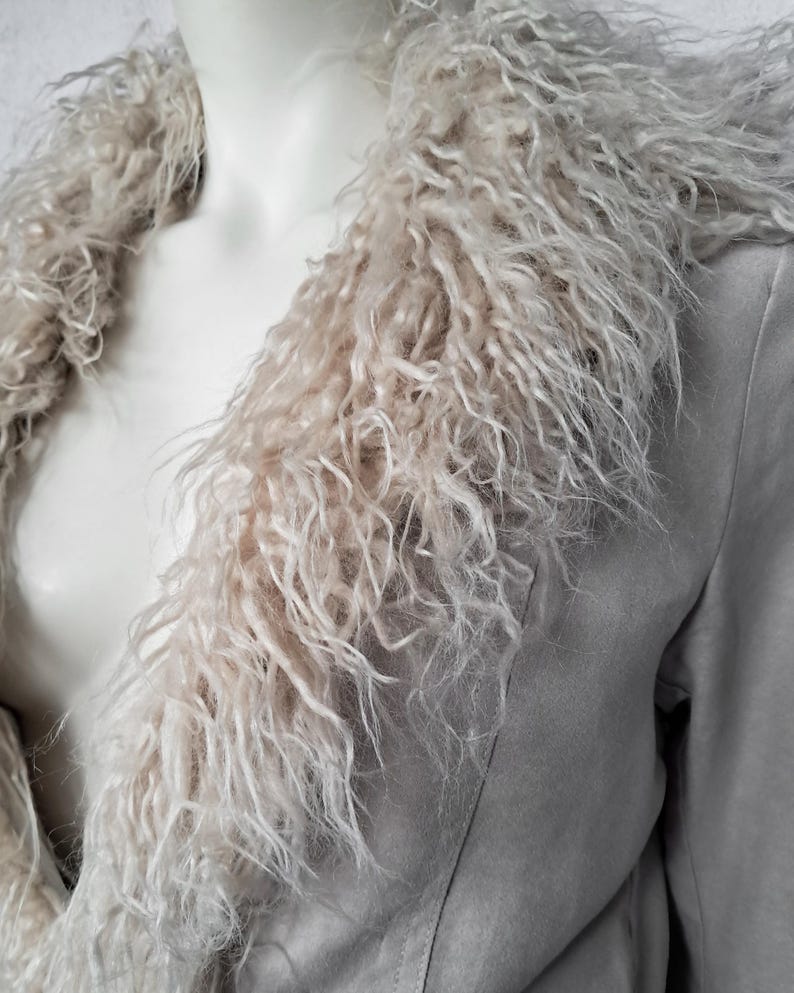May include: A light gray faux suede jacket with a shaggy, light beige faux fur collar. The jacket has a classic lapel design and a soft, textured appearance. The faux fur collar adds a touch of luxury and warmth to the garment.