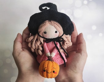 May include: A crocheted witch doll with a black hat, pink dress, and long brown hair. The doll is holding a small orange pumpkin with a smiling face.