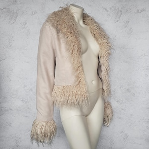 May include: A light beige jacket with faux fur trim on the collar, cuffs, and hem. The jacket is displayed on a mannequin. The jacket has long sleeves and a cropped length. The faux fur is a lighter shade of beige.