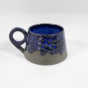 Könnte beinhalten: Eine handgefertigte Keramiktasse mit dunkelblauem Innenraum und strukturierter Außenseite. Die Tasse hat einen dunkelblauen Henkel und einen grauen Boden. Die Außenseite hat ein gesprenkeltes Muster mit blauen und violetten Akzenten. Die Tasse ist mit dem Text "HAD <<KRAMIK" versehen.