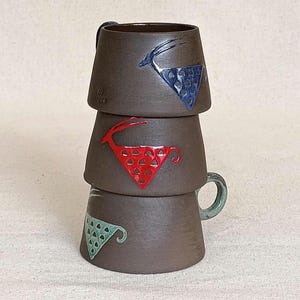 May include: Three stacked brown ceramic mugs with colorful geometric designs. The top mug has a blue triangle with a deer silhouette. The middle mug has a red triangle with a deer. The bottom mug has a green triangle.