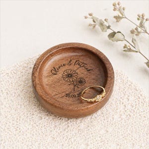 May include: A round, wooden ring dish with engraved floral design and the names "Olivia & Patrick" along with the date "10.09.20". A gold ring with small stones rests inside the dish. The dish is on a white lace surface.