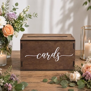 Puede incluir: Una caja rectangular de madera de color marrón oscuro con una ranura horizontal en la tapa superior. El panel frontal presenta la palabra cards escrita en una fuente cursiva blanca. La caja está construida con juntas de esquina visibles.