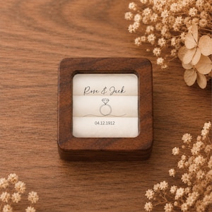 May include: A square dark brown wooden ring box with a clear lid. The interior features a white velvet insert with the printed text Rose & Jack, a line drawing of a diamond ring, and the date 04.12.1912 centered on the white fabric surface.