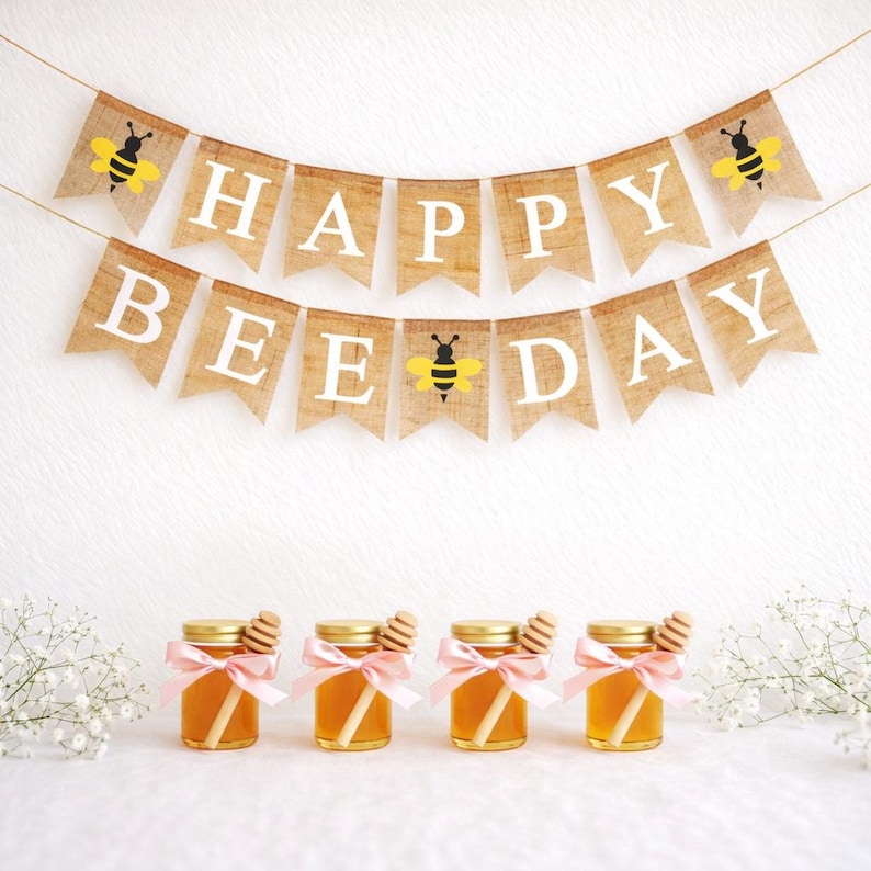 May include: A banner that reads "HAPPY BEE DAY" with bee illustrations, above four small jars of honey tied with pink ribbons and honey dippers. The jars have gold lids and are filled with golden honey.