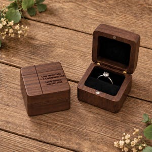 Peut inclure: Une boîte à bague en bois brun foncé avec un couvercle à charnière et une doublure intérieure noire. Le couvercle est gravé avec le texte : Love Never Fails — 1 Corinthians 13:8, Lee & Gemma, July 28th 2026. Une bague argentée avec une pierre ronde.