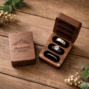 May include: A wooden ring box with three ring slots, holding a gold band, a diamond engagement ring, and a diamond band. The box is open, revealing a black velvet interior. The lid is closed, engraved with a mountain scene and the names "Olivia & Patrick" and the date "10.09.2026".