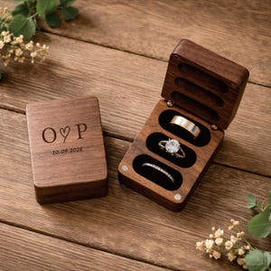 May include: A wooden ring box with three slots, one open and holding three rings. The closed box has the initials "O♡P" and the date "10.09.2026" engraved on the lid. The rings include a diamond engagement ring and a gold band.