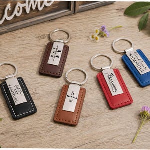 May include: Five rectangular keychains in black, dark brown, light brown, red, and blue, each featuring a silver metal plate with custom engravings including names like SAMMUEL, dates, and phrases like BEST DAD, attached to a silver metal ring.