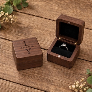 Puede incluir: Dos cajas cuadradas para anillos de madera oscura con cierres magnéticos. Una caja está cerrada y grabada con las letras L y Y y la fecha 6.9.20xx. La otra caja está abierta, revelando un interior de terciopelo negro con un anillo de plata.
