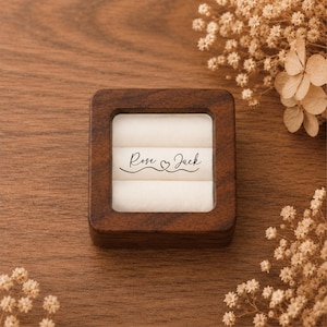 May include: A square dark brown wooden ring box with a clear lid and a white velvet interior. The center of the white velvet features the black printed text "Rose" followed by a small heart icon and the name "Jack" in a cursive script font.