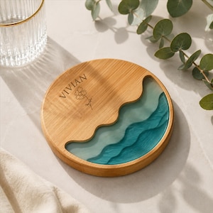 Puede incluir: Un posavasos redondo de bambú con acabado de veta de madera natural en un lado y una incrustación de resina ondulada en tonos azul verdoso y azul claro en el otro. El nombre VIVIAN está grabado sobre una pequeña ilustración lineal de una flor.