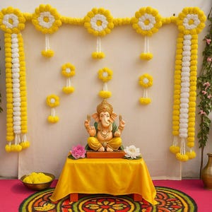 May include: A colourful display featuring a golden statue of Ganesha on a yellow table, adorned with pink and white flowers. The backdrop includes yellow and white floral garlands, with decorative vases and a basket of coconuts, creating a festive atmosphere.