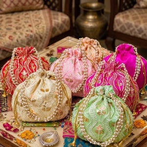 Può includere: Una collezione di borse ricamate con coulisse in vari colori, tra cui rosso, rosa e verde. Ogni borsa è adornata con motivi intricati e impreziosita da perline simili a perle. Le borse sono disposte su una superficie decorativa.