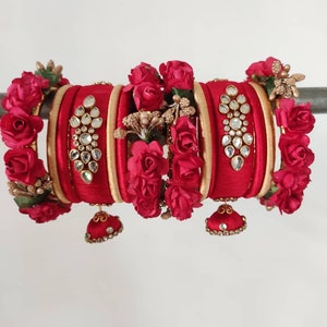 May include: A set of red silk thread bangles with gold accents and jeweled embellishments. The bangles are decorated with red fabric roses and green leaves.