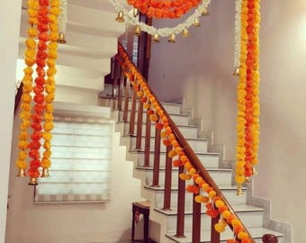 May include: A staircase decorated with garlands of orange, yellow, and white flowers. Golden bells hang from the garlands. The wooden handrail is also adorned with orange and yellow flower garlands. The stairs are made of white marble.