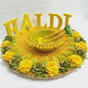 Op de afbeelding: Een decoratief geel Haldi-dienblad met een centrale kom, twee stokjes en het woord "HALDI" in grote gele letters. De lade is versierd met gele en groene rozen en een gouden rand, wat zorgt voor een feestelijke en kleurrijke presentatie.