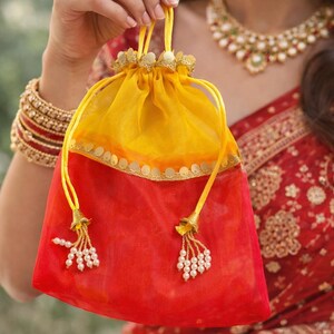 Può includere: Un'accattivante pochette bicolore con coulisse rossa e gialla. La pochette presenta abbellimenti color oro sulla parte superiore e alle estremità dei cordini, adornati con piccole perle bianche. La pochette è tenuta da una persona.