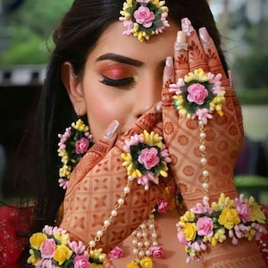 May include: A bridal jewellery set featuring pink and yellow flowers, pearls, and a delicate chain. The set includes a maang tikka, earrings, bangles, and rings.