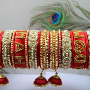 May include: A collection of red and gold bangles with intricate beadwork and embellishments. Some bangles feature the names "MAH" and "DOM" in gold lettering. Each bangle has a small, red, bell-shaped charm hanging from it.