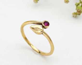 May include: A gold ring with a leaf design and a small red gemstone. The ring is open and can be adjusted to fit different finger sizes.