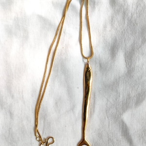 Op de afbeelding: Een gouden ketting met een lange, dunne, gouden vis hanger.