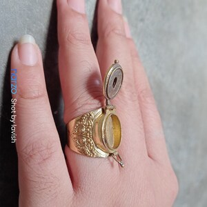 Puede incluir: Un anillo de oro con un compartimento de forma ovalada que se abre. El compartimento est&aacute; abierto y el anillo se lleva en un dedo.
