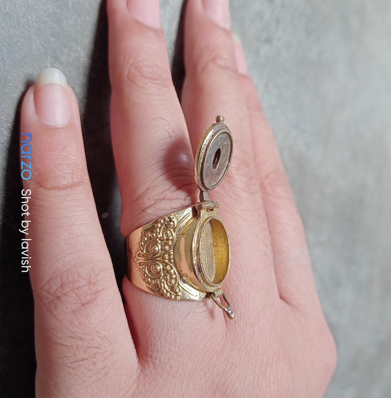 Puede incluir: Un anillo de oro con un compartimento de forma ovalada que se abre. El compartimento est&aacute; abierto y el anillo se lleva en un dedo.
