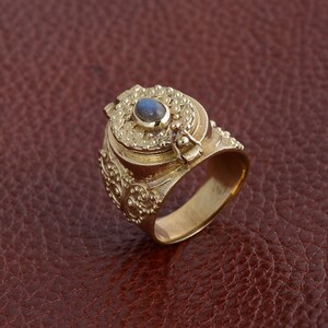 Puede incluir: Un anillo de oro con un compartimento con bisagras y una piedra preciosa azul engastada en la parte superior. El anillo est&aacute; intrincadamente detallado con patrones florales.